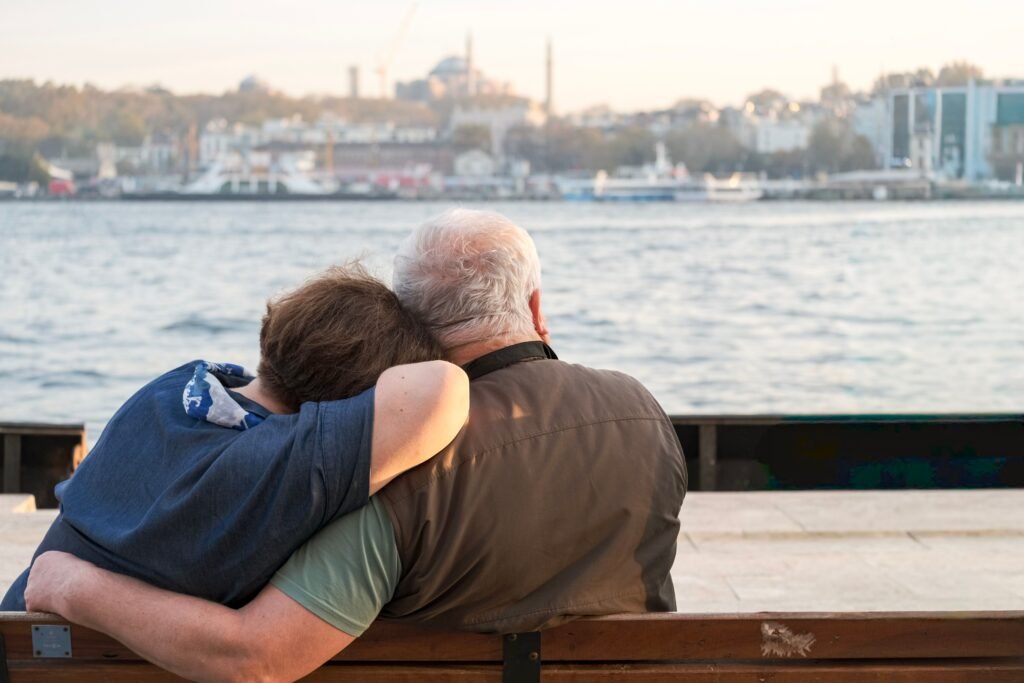 An elderly man and younger person embracing while looking at the water, symbolizing the lasting rewards of a longevity mindset
