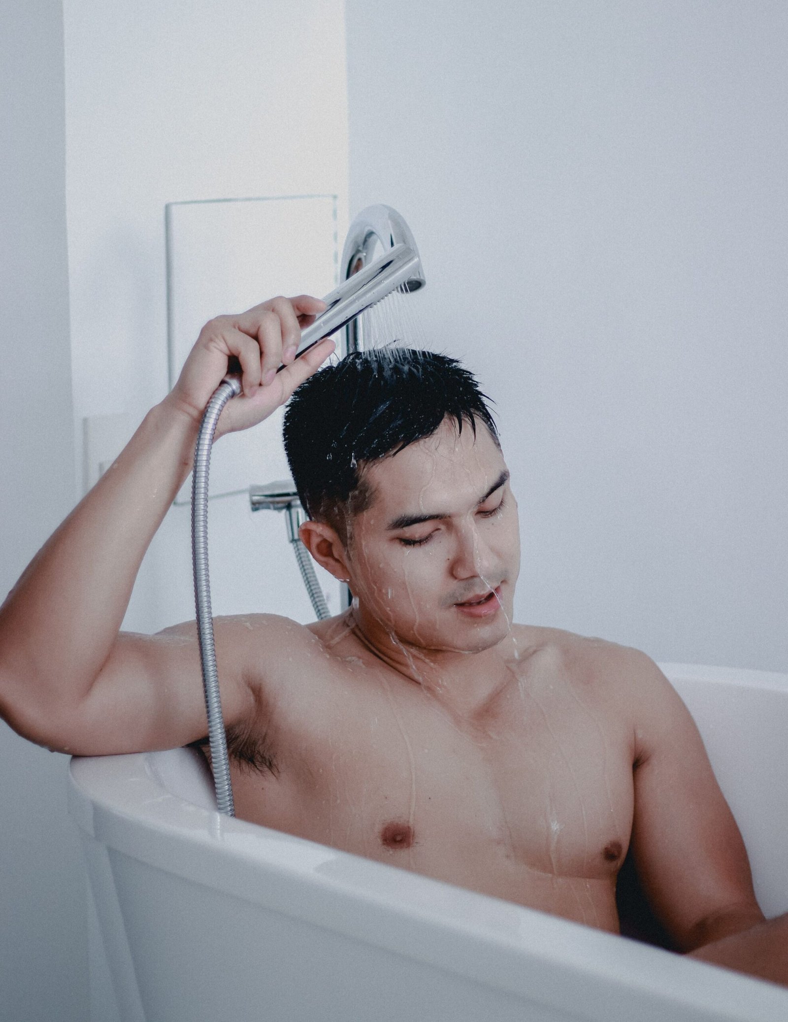 Man holding a handheld shower head over his head while sitting in a bathtub