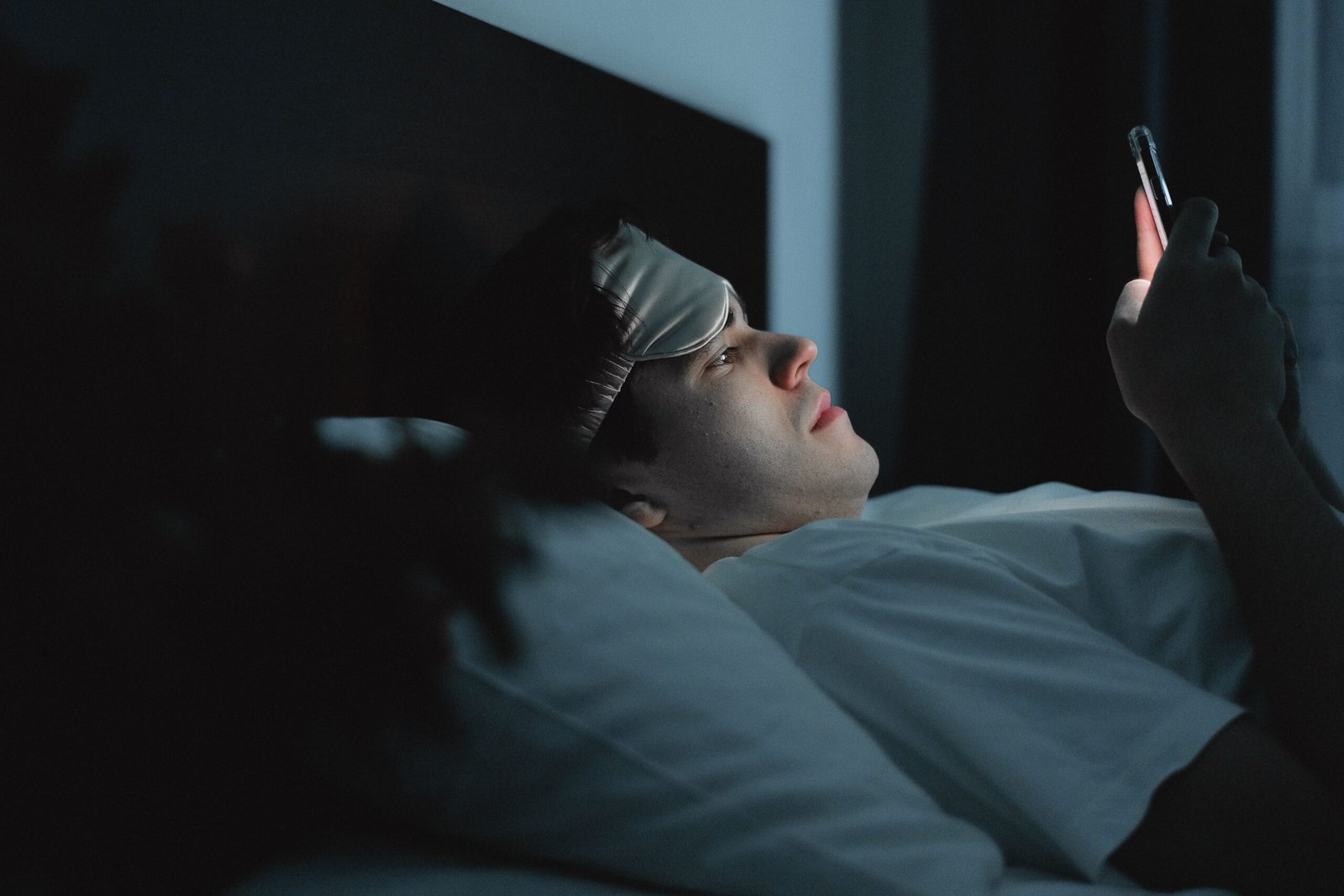 Man lying in bed at night scrolling his phone with a sleep mask on his forehead, illustrating the habit of scrolling before sleep